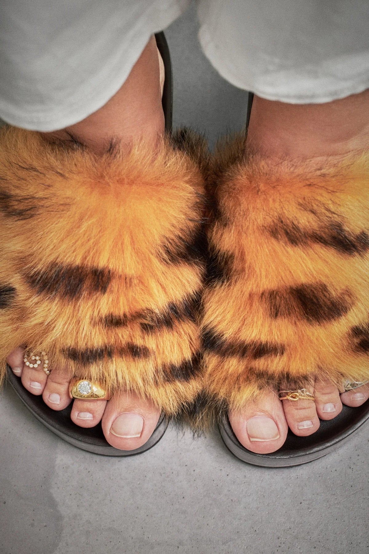 Feet wearing fluffy orange and black slides with meaningful everyday jewelry on toes, emphasizing personal style.