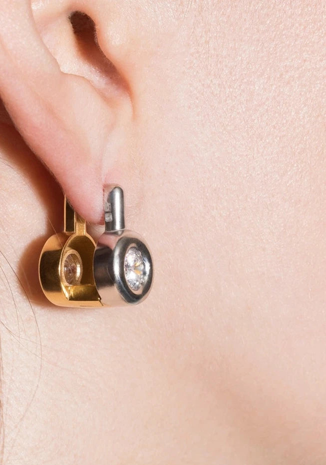 A close-up of a model's ear wearing a pair of gold and silver geometric stud earrings with clear gemstones.