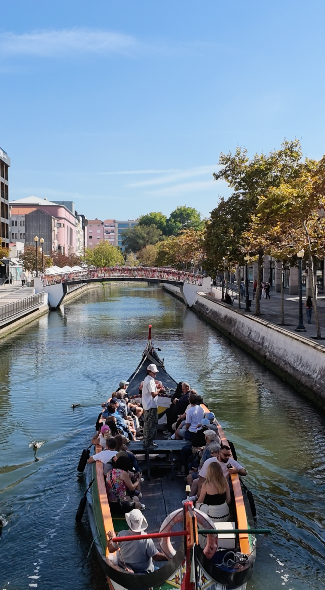 A gondola boat with passengers travels down a canal in an urban setting, with a red bridge, trees, and buildings visible in the background under a clear blue sky.