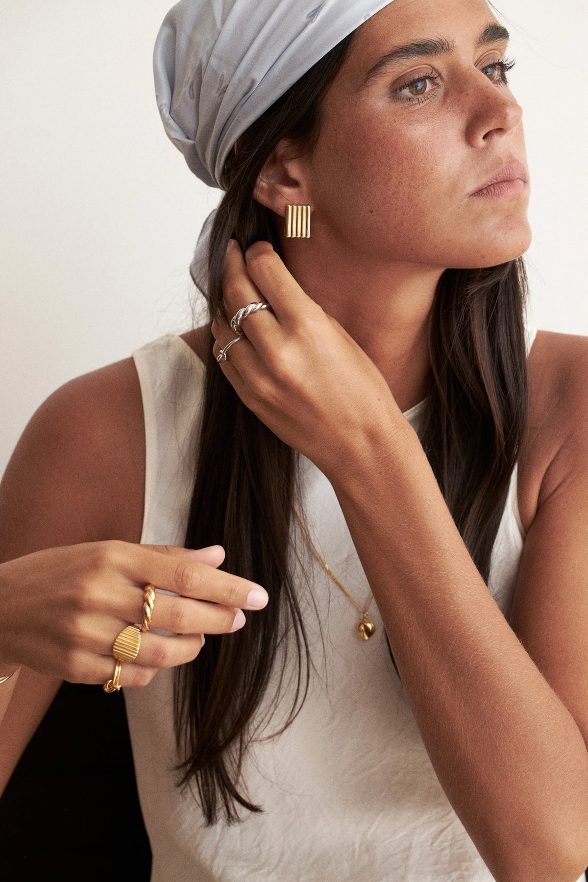 Cannelé Rectangular Ribbed Earrings. A woman wearing a white sleeveless top, a light gray headscarf, and multiple gold rings, with one gold ring with vertical bars shown on her finger. She is looking to the side against a plain light background. where can I find cool earrings online?