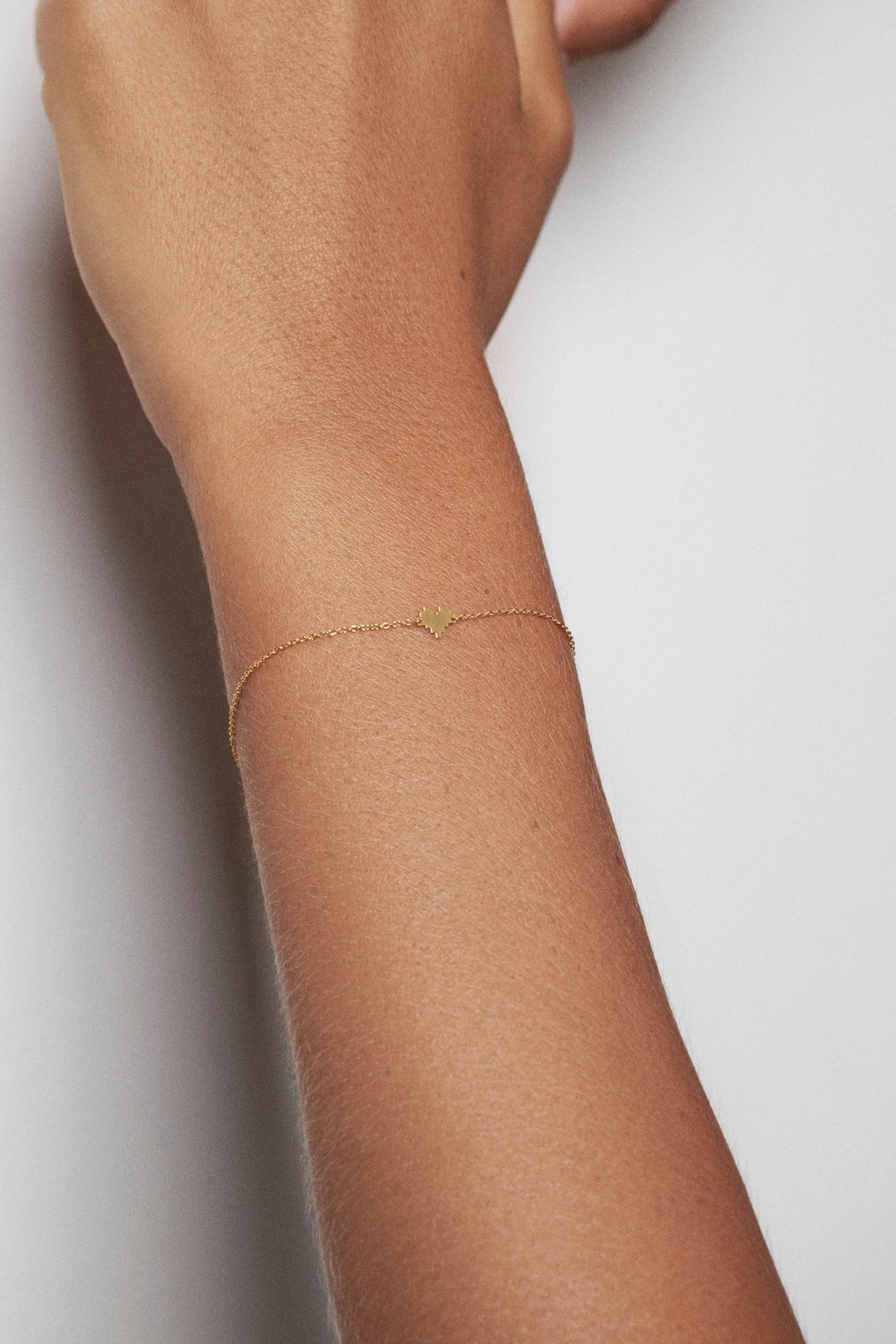 A delicate gold chain bracelet with a small, minimalist gold charm worn on a person's wrist, shown against a clean, light gray background.