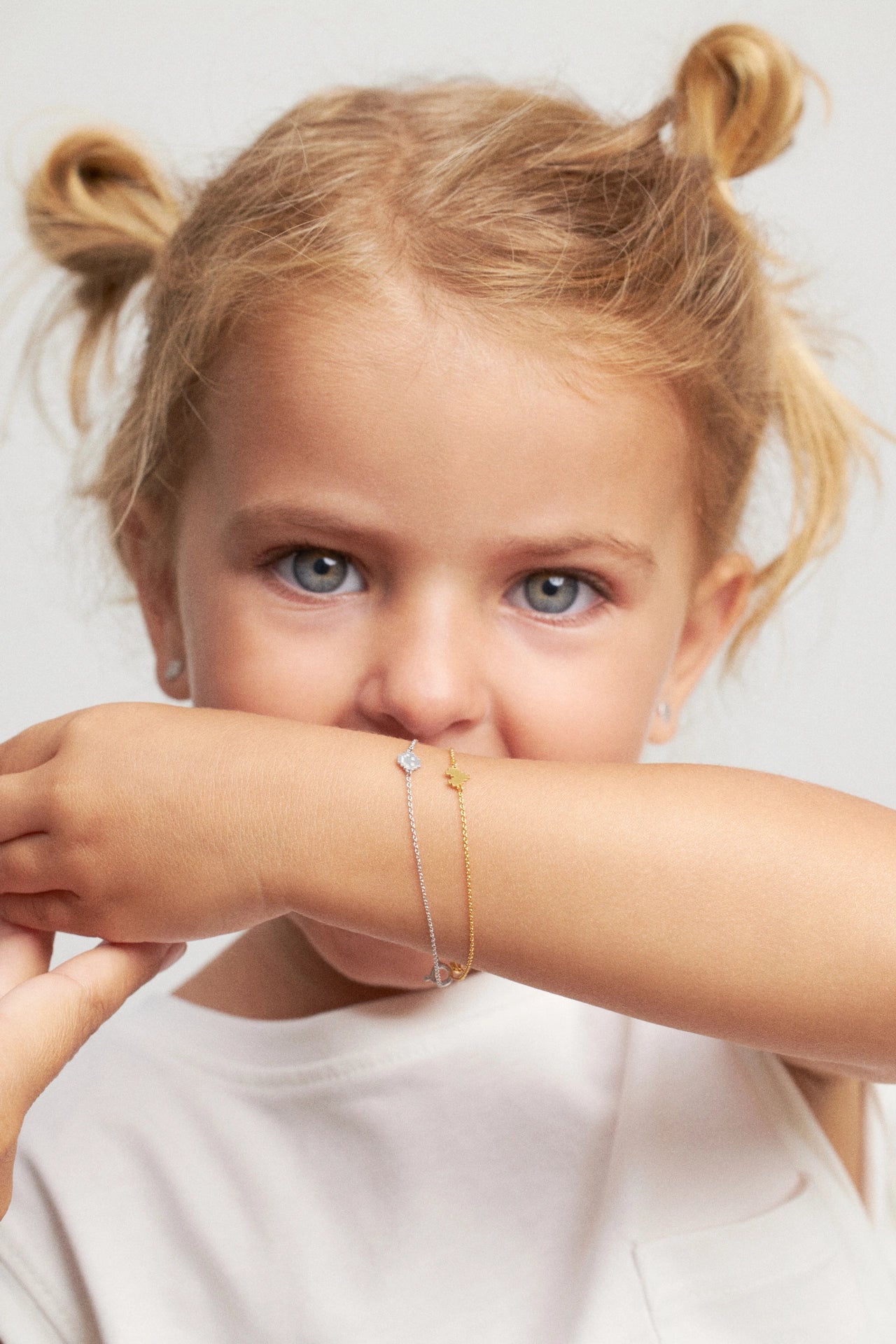 A close-up lifestyle shot of a young girl with blonde pigtails wearing delicate gold and silver chain bracelets on her wrist, holding her arm up to show the jewelry against a light gray background.