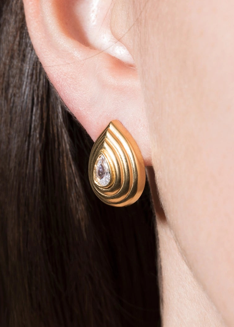 A close-up of a gold teardrop-shaped earring with concentric ridges and a small clear central stone, worn on a person's ear.
